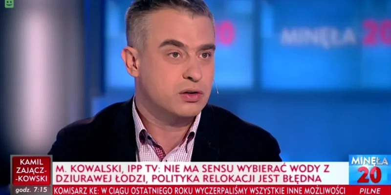 Mężczyzna z mikrofonem w uchu przemawia w studiu telewizyjnym podczas programu informacyjnego, a na pasku pod zdjęciem widoczny jest tekst o zobowiązaniach Polski w sprawie uchodźców.