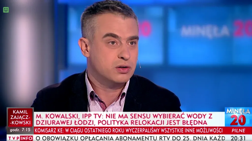 Mężczyzna z mikrofonem w uchu przemawia w studiu telewizyjnym podczas programu informacyjnego, a na pasku pod zdjęciem widoczny jest tekst o zobowiązaniach Polski w sprawie uchodźców.