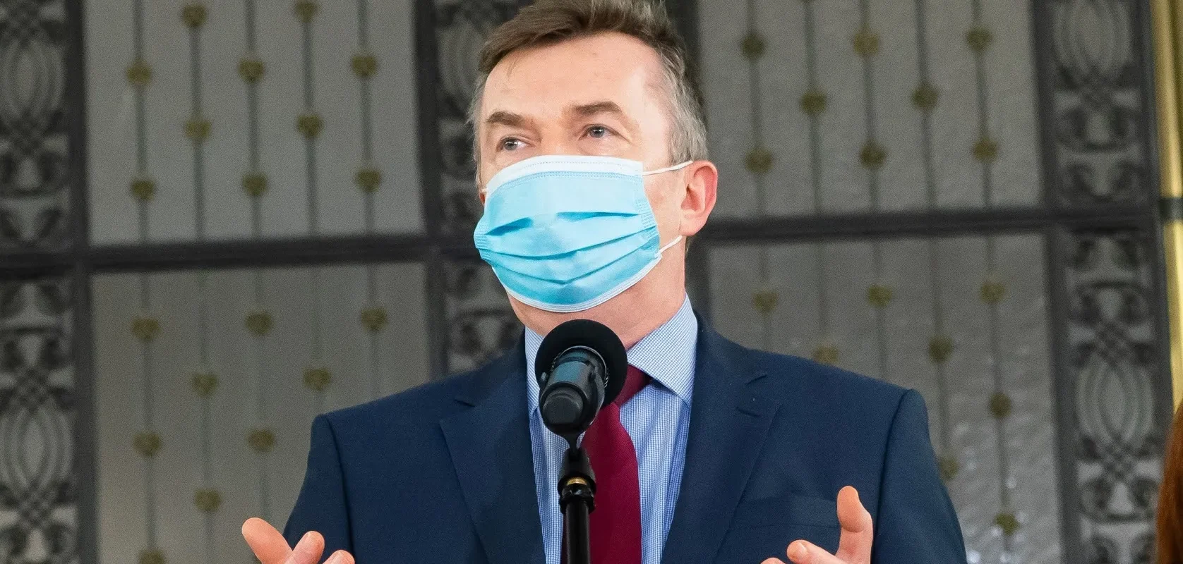 Mężczyzna w garniturze i niebieskiej maseczce ochronnej stoi przy mikrofonie i gestykuluje podczas przemówienia.