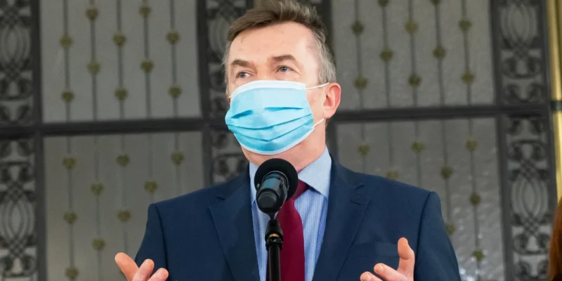 Mężczyzna w garniturze i niebieskiej maseczce ochronnej stoi przy mikrofonie i gestykuluje podczas przemówienia.