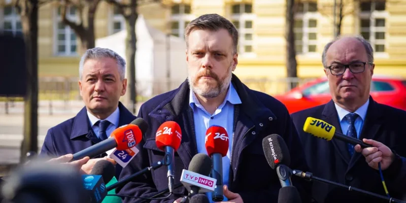 Politycy Lewicy stoją na konferencji prasowej w sprawie Ukrainy, otoczeni mikrofonami dziennikarzy, na tle żółtej kamienicy.