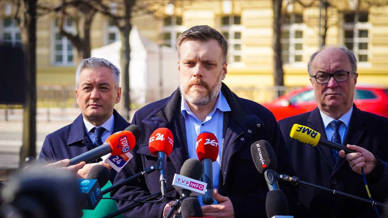 Politycy Lewicy stoją na konferencji prasowej w sprawie Ukrainy, otoczeni mikrofonami dziennikarzy, na tle żółtej kamienicy.