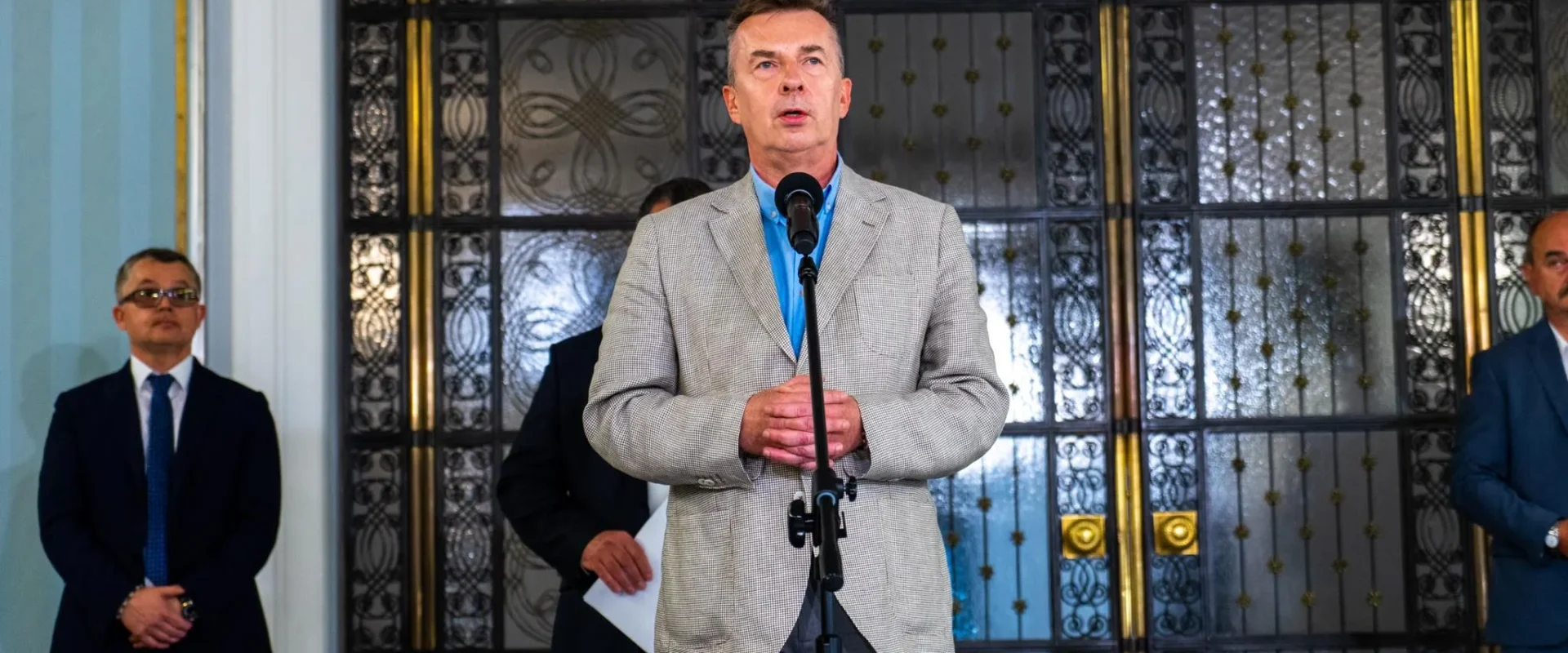 Mężczyzna w jasnoszarym żakiecie i niebieskiej koszuli stoi przy mikrofonie podczas konferencji prasowej, otoczony innymi mężczyznami w garniturach, a w tle widoczne ozdobne drzwi.
