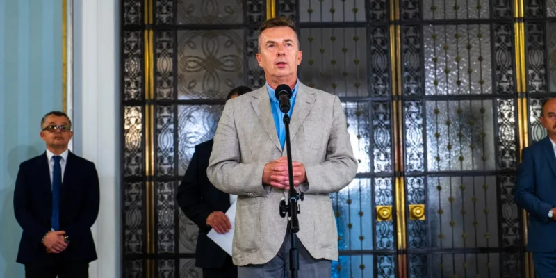 Mężczyzna w jasnoszarym żakiecie i niebieskiej koszuli stoi przy mikrofonie podczas konferencji prasowej, otoczony innymi mężczyznami w garniturach, a w tle widoczne ozdobne drzwi.