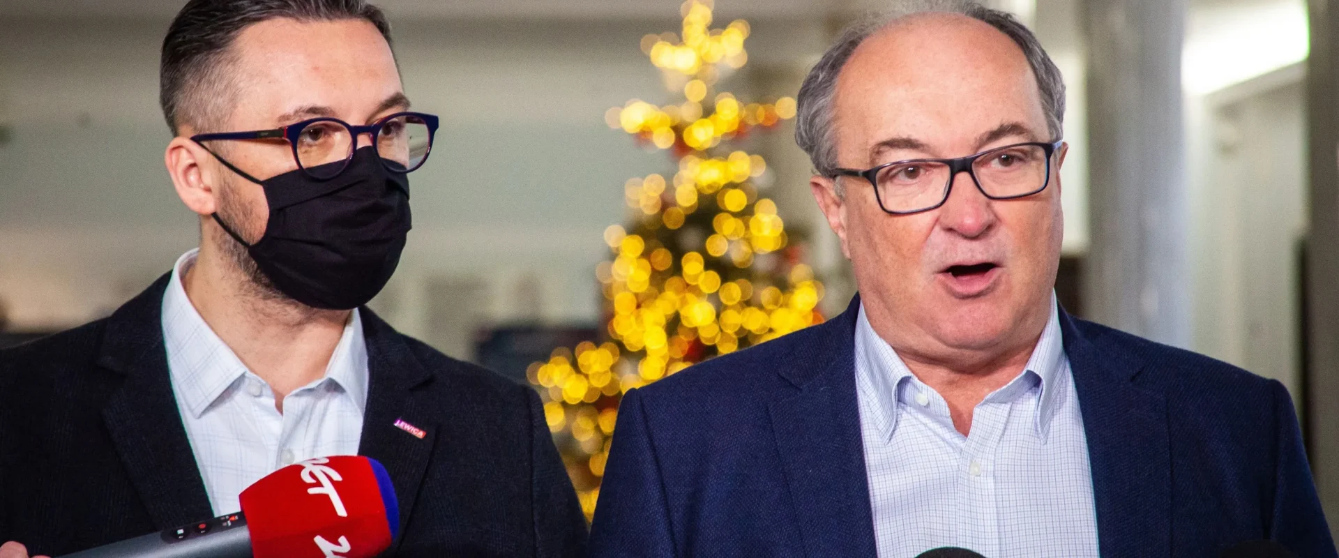 Dwóch mężczyzn w garniturach na konferencji prasowej stojących obok siebie przy licznych mikrofonach, z których jeden ma założoną czarną maseczkę, a w tle widać migoczącą choinkę.