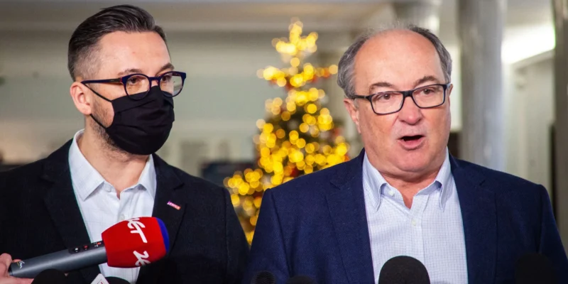 Dwóch mężczyzn w garniturach na konferencji prasowej stojących obok siebie przy licznych mikrofonach, z których jeden ma założoną czarną maseczkę, a w tle widać migoczącą choinkę.