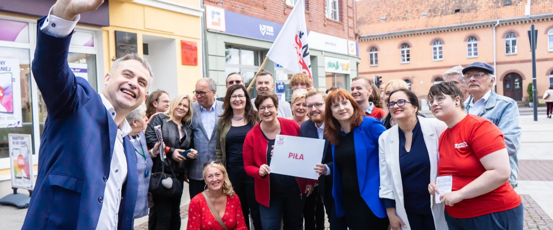 Na zdjęciu grupa ludzi stoi na ulicy, pozując do selfie z uśmiechami, trzymając tabliczkę z napisem "Piła".