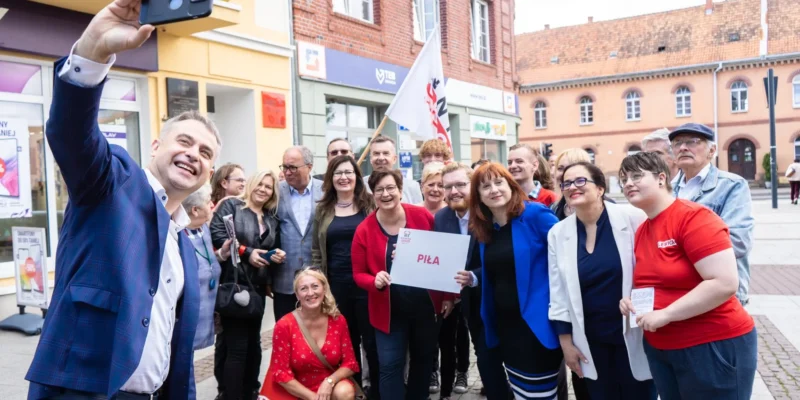 Na zdjęciu grupa ludzi stoi na ulicy, pozując do selfie z uśmiechami, trzymając tabliczkę z napisem "Piła".