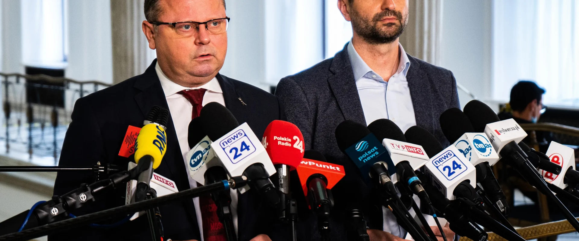 Dwóch mężczyzn w garniturach stoi przy licznym zestawie mikrofonów podczas konferencji prasowej.