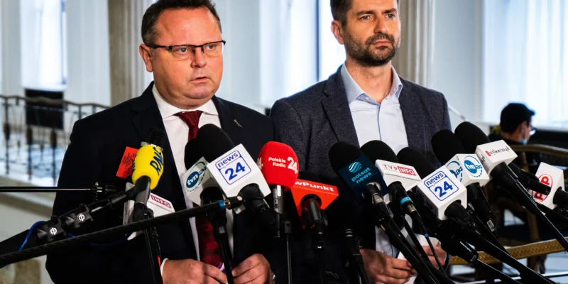Dwóch mężczyzn w garniturach stoi przy licznym zestawie mikrofonów podczas konferencji prasowej.