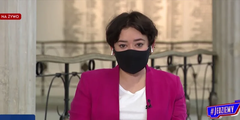 Kobieta prowadząca program telewizyjny na żywo, ubrana w różowy żakiet i czarną maseczkę, siedzi w studiu przed mikrofonem.