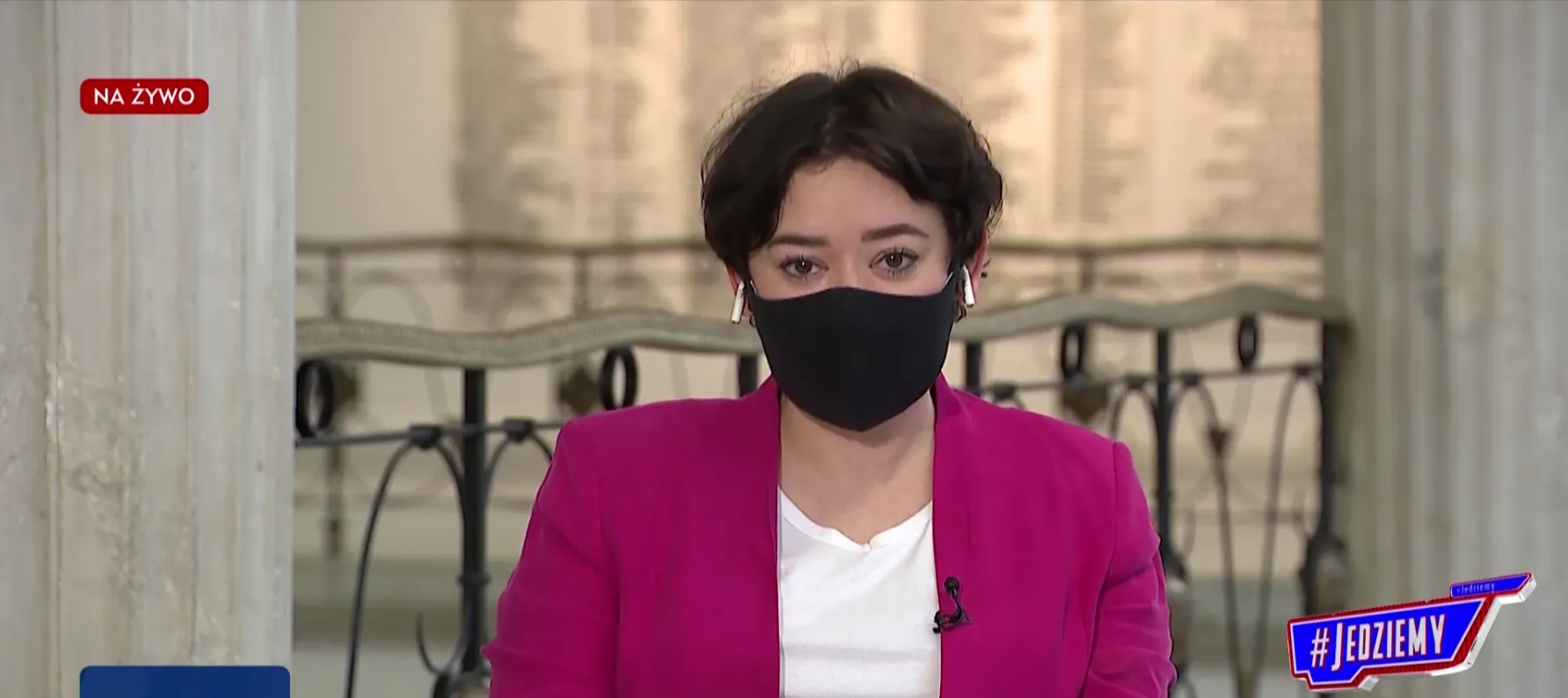 Kobieta prowadząca program telewizyjny na żywo, ubrana w różowy żakiet i czarną maseczkę, siedzi w studiu przed mikrofonem.