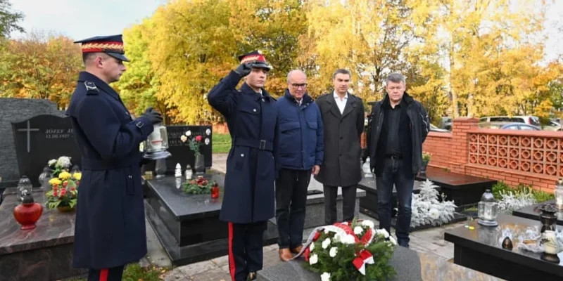 Na cmentarzu w jesiennej scenerii grupa ludzi wraz z dwoma żołnierzami w mundurach salutuje przy grobie ozdobionym wieńcem kwiatów — Pamiętamy.