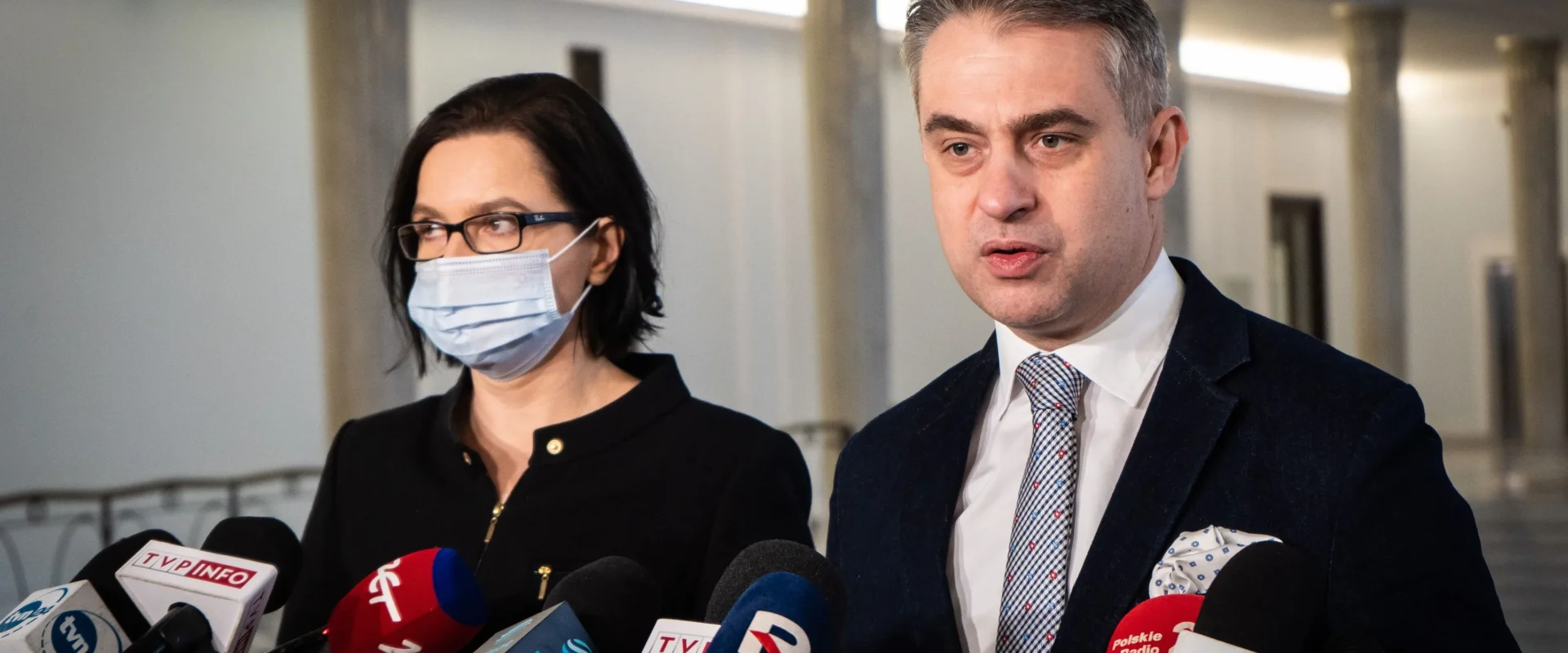 Dwóch polityków przy licznych mikrofonach mediów podczas konferencji prasowej w sprawie kar za Turów; mężczyzna w garniturze zabiera głos, obok stoi kobieta w maseczce.