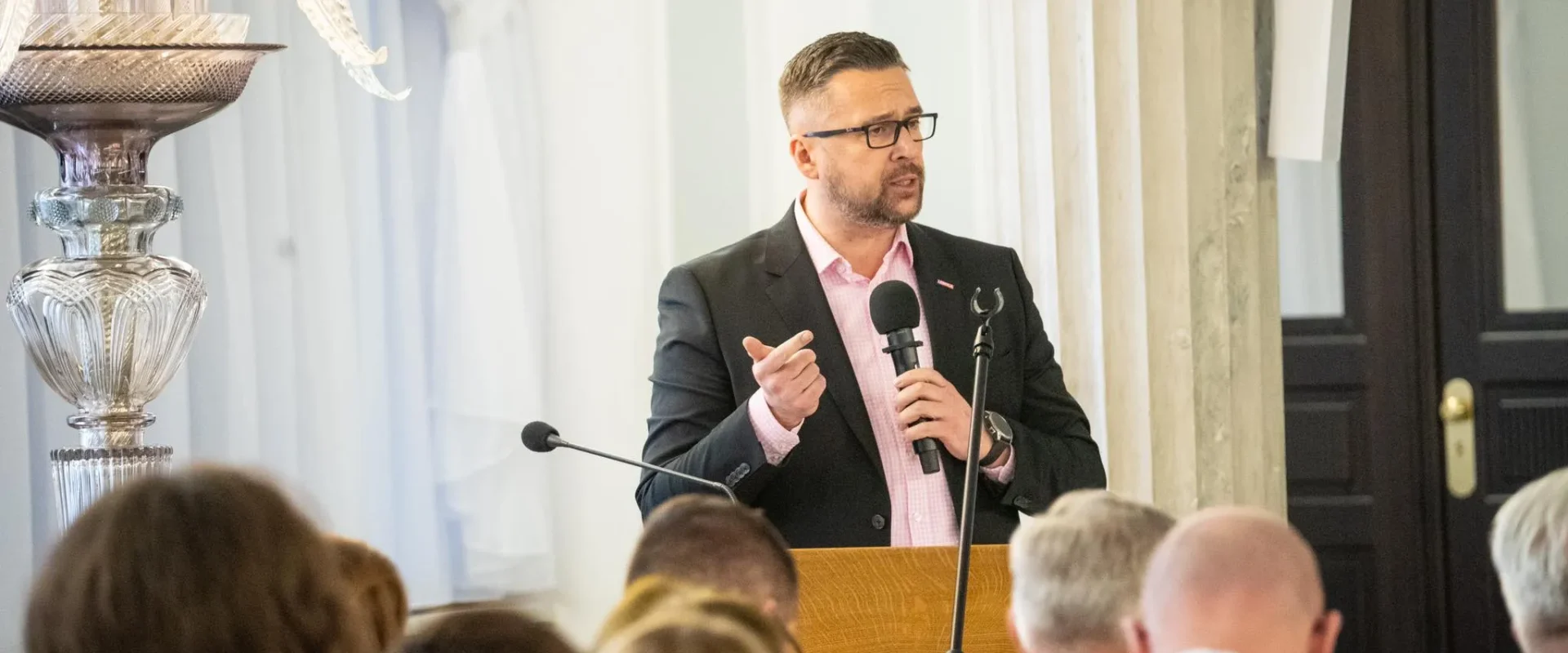 Mężczyzna w garniturze i różowej koszuli przemawia z mikrofonem przy mównicy podczas spotkania, w tle widać słuchaczy i ozdobny żyrandol.