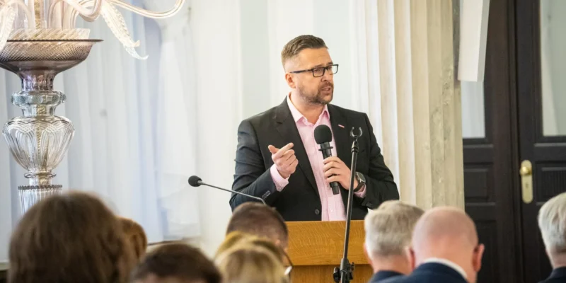 Mężczyzna w garniturze i różowej koszuli przemawia z mikrofonem przy mównicy podczas spotkania, w tle widać słuchaczy i ozdobny żyrandol.