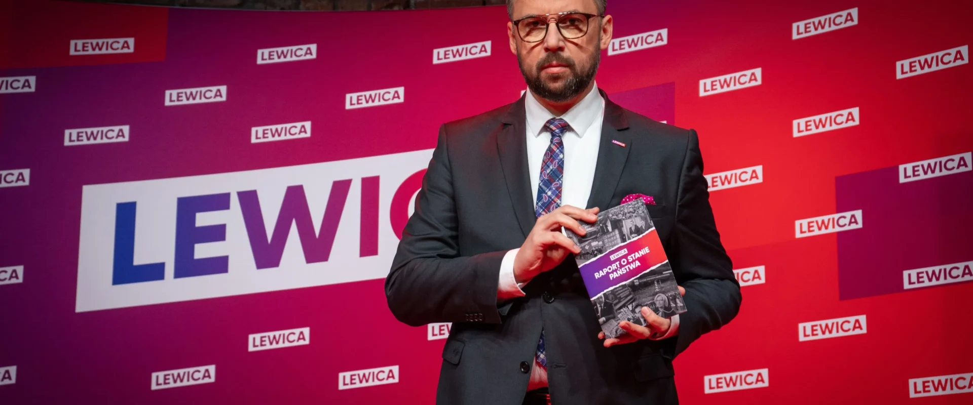 Mężczyzna w eleganckim garniturze prezentuje książkę zatytułowaną "Raport o stanie państwa" na tle banera z logo Lewicy.