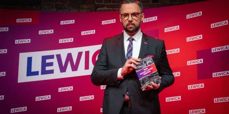 Mężczyzna w eleganckim garniturze prezentuje książkę zatytułowaną "Raport o stanie państwa" na tle banera z logo Lewicy.