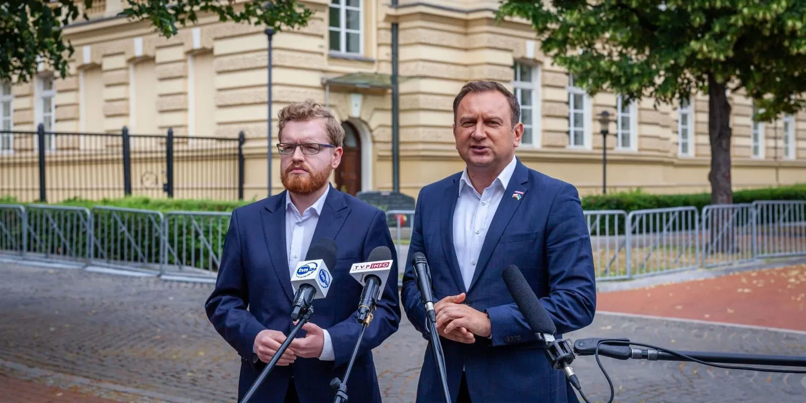 Dwóch mężczyzn w garniturach stoi przed mikrofonami na konferencji prasowej na zewnątrz, przed żółtawym budynkiem oraz barierkami.