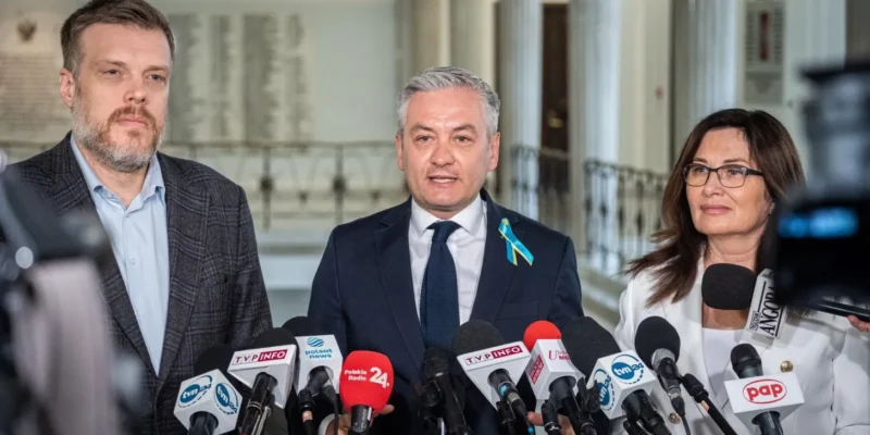 Na zdjęciu widać trzech polityków podczas konferencji prasowej, otoczonych mikrofonami, w formalnym otoczeniu.