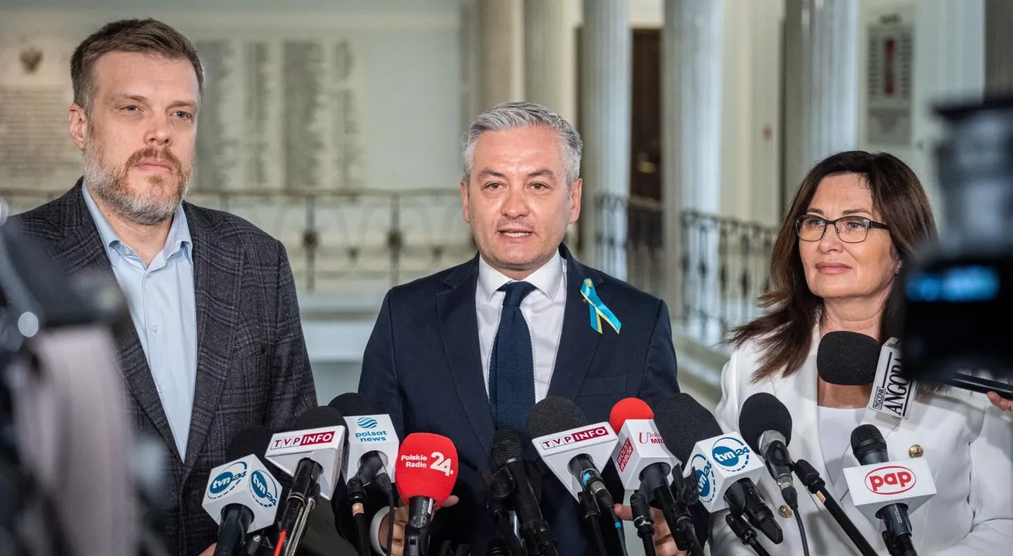 Na zdjęciu widać trzech polityków podczas konferencji prasowej, otoczonych mikrofonami, w formalnym otoczeniu.