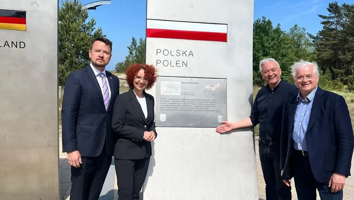 Czterech dorosłych mężczyzn w garniturach stoi obok granicznego pomnika z napisem Polska Polen, na tle zieleni.