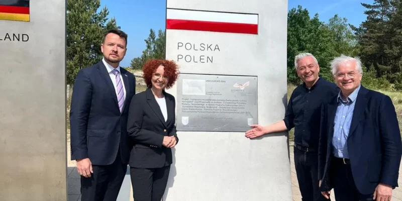 Czterech dorosłych mężczyzn w garniturach stoi obok granicznego pomnika z napisem Polska Polen, na tle zieleni.