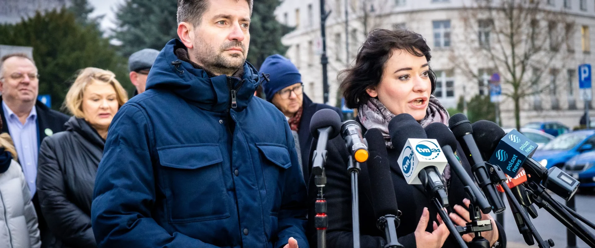 Mężczyzna i kobieta zabierają głos podczas konferencji prasowej o praworządności, stojąc przy zestawie mikrofonów, otoczeni przez innych uczestników i budynki w tle.