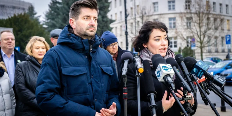 Mężczyzna i kobieta zabierają głos podczas konferencji prasowej o praworządności, stojąc przy zestawie mikrofonów, otoczeni przez innych uczestników i budynki w tle.