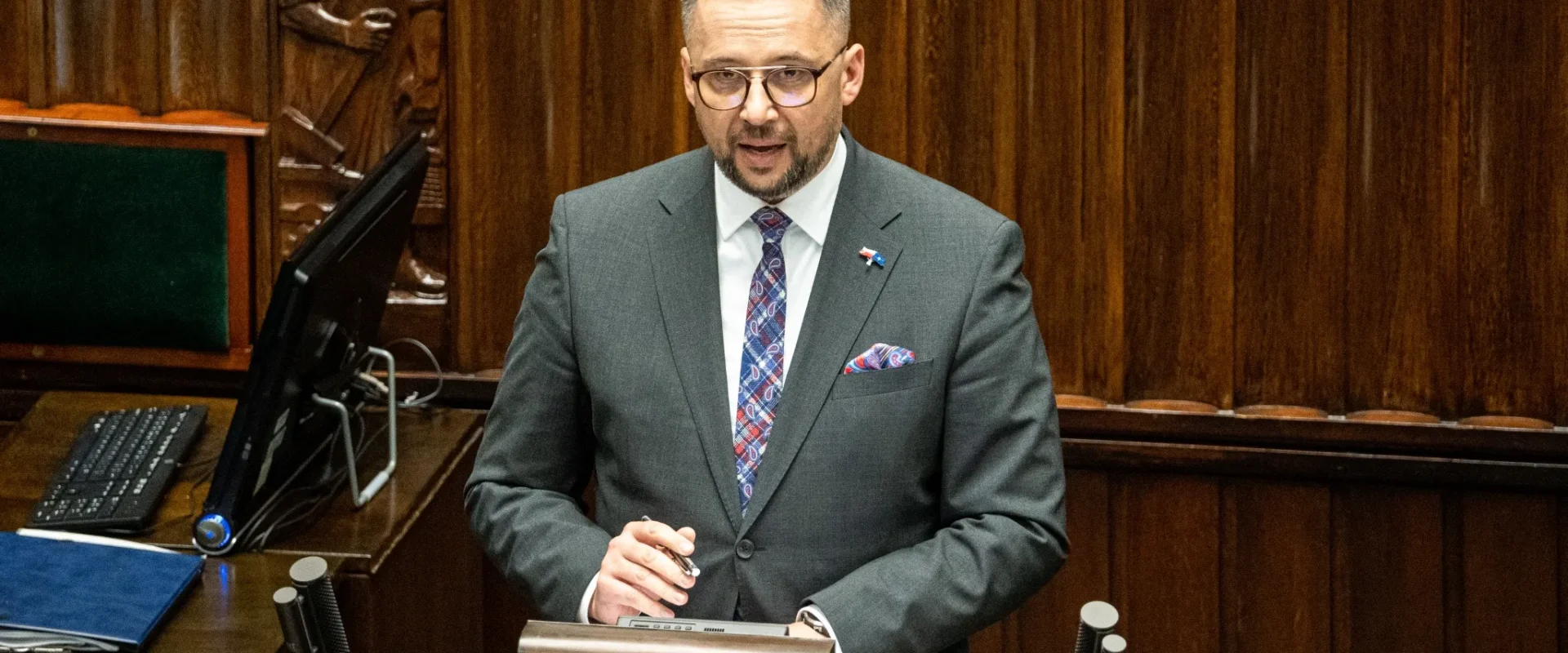 Mężczyzna w smokingu, stojący przy mównicy w sali obrad, mówi do zgromadzenia.