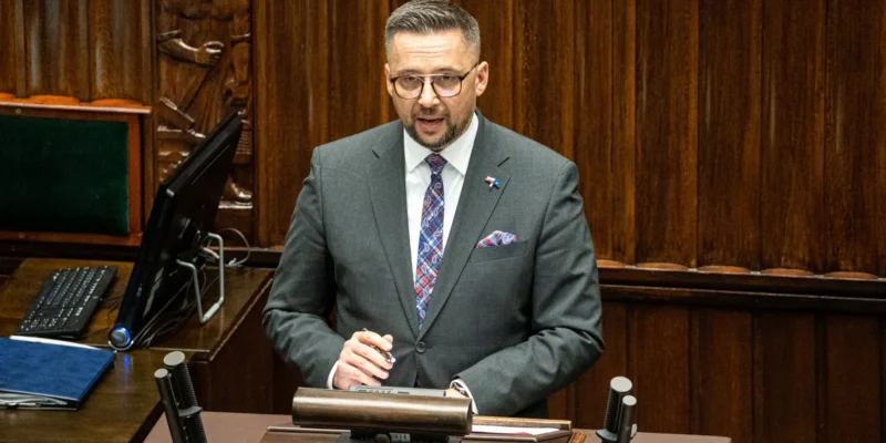 Mężczyzna w smokingu, stojący przy mównicy w sali obrad, mówi do zgromadzenia.