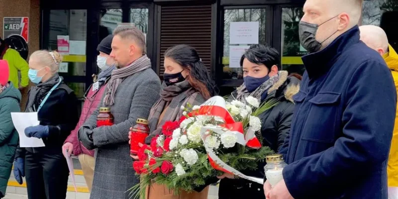 Grupa ludzi stoi z kwiatami i zniczami w rękach podczas ceremonii upamiętniającej, ubrana w maski ochronne w miejscu publicznym.