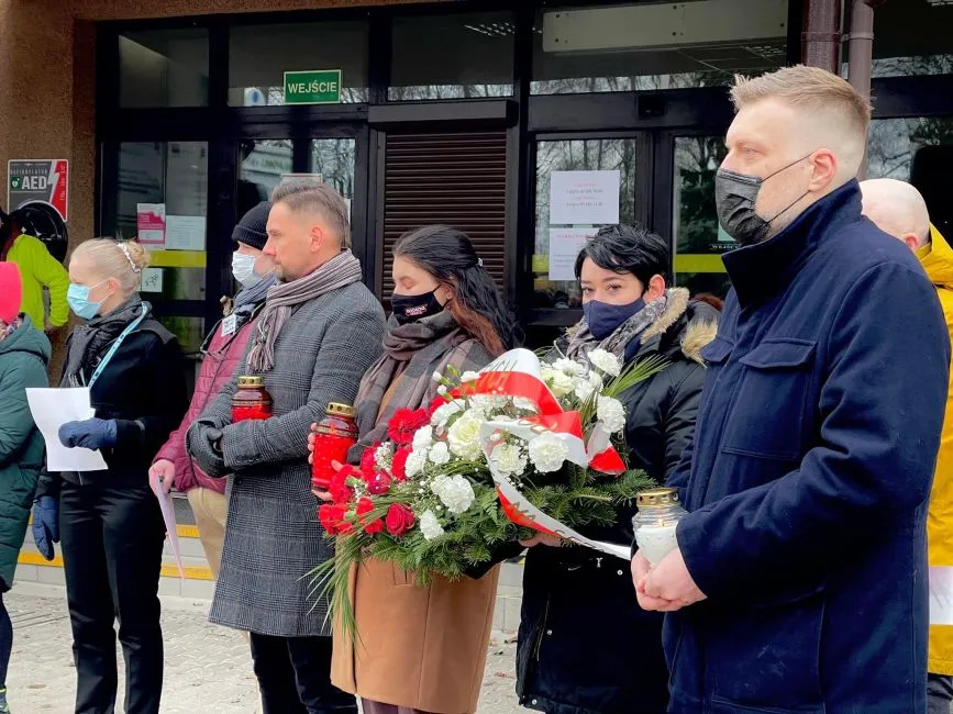 Grupa ludzi stoi z kwiatami i zniczami w rękach podczas ceremonii upamiętniającej, ubrana w maski ochronne w miejscu publicznym.