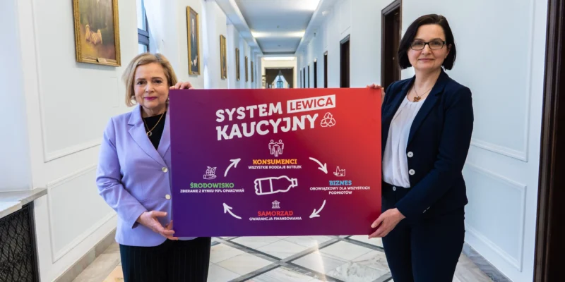 Dwie kobiety w eleganckich ubraniach stoją w korytarzu i trzymają duży plakat z napisem System Kaucyjny Lewica.