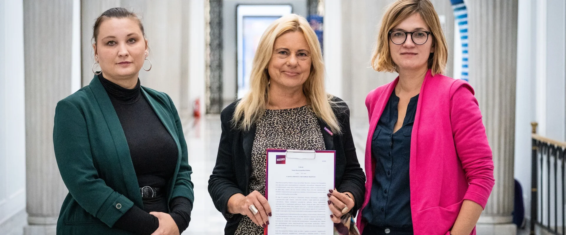 Trzy kobiety stoją w eleganckim korytarzu z kolumnami; pośrodku blondynka trzyma plik z dokumentem, a po bokach stoją w zielonej i różowej marynarce.