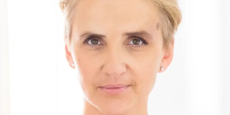 Portret kobiety o krótkich blond włosach w czerwonym topie, patrzącej w kamerę na jasnym tle.