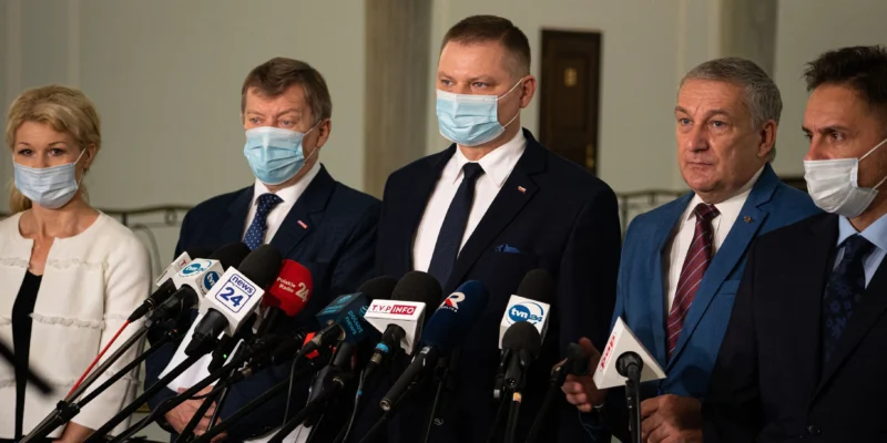 Grupa mężczyzn i kobiet w maseczkach stoi przy mikrofonach podczas konferencji prasowej w budynku administracyjnym.