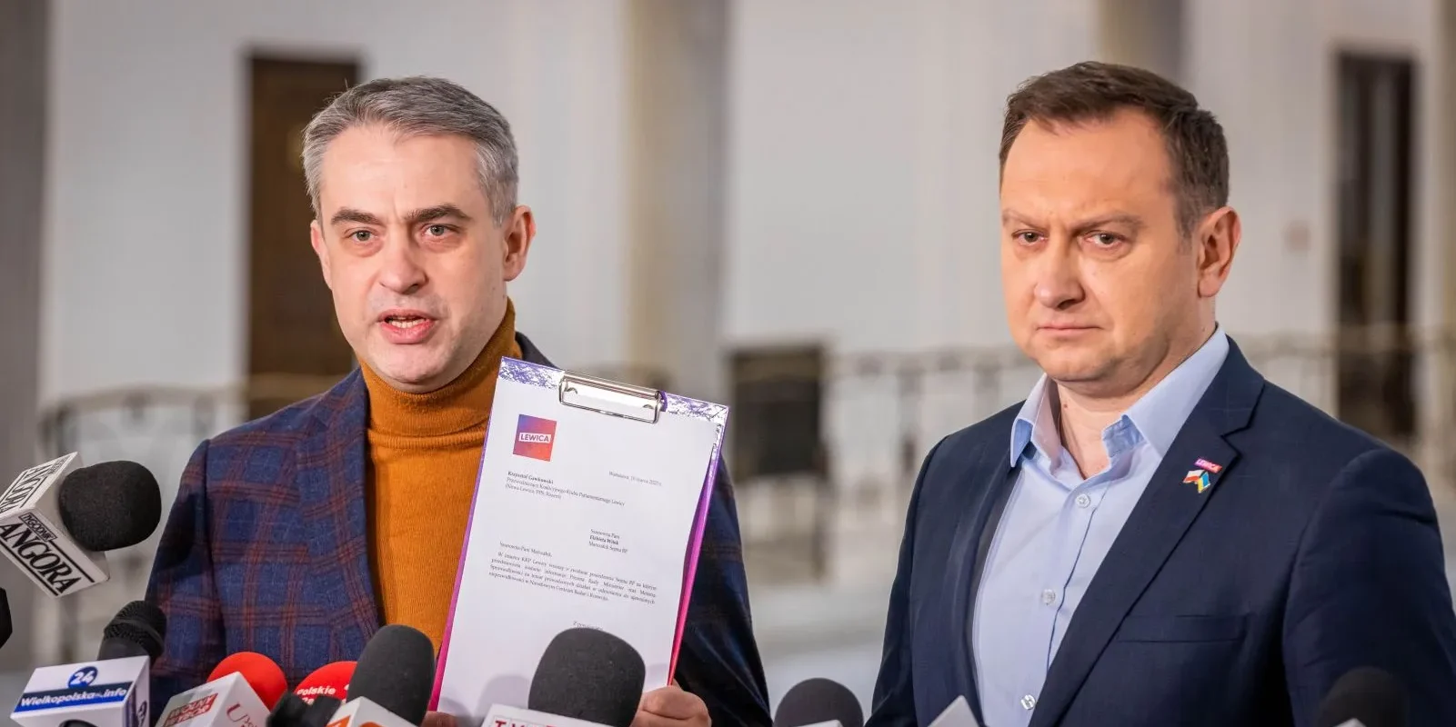 Dwóch mężczyzn w garniturach na konferencji prasowej, jeden trzyma klipboard z dokumentem, otoczeni licznymi mikrofonami.