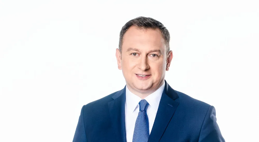 Portret mężczyzny w granatowym garniturze i niebieskim krawacie, uśmiechnięty, na białym tle.