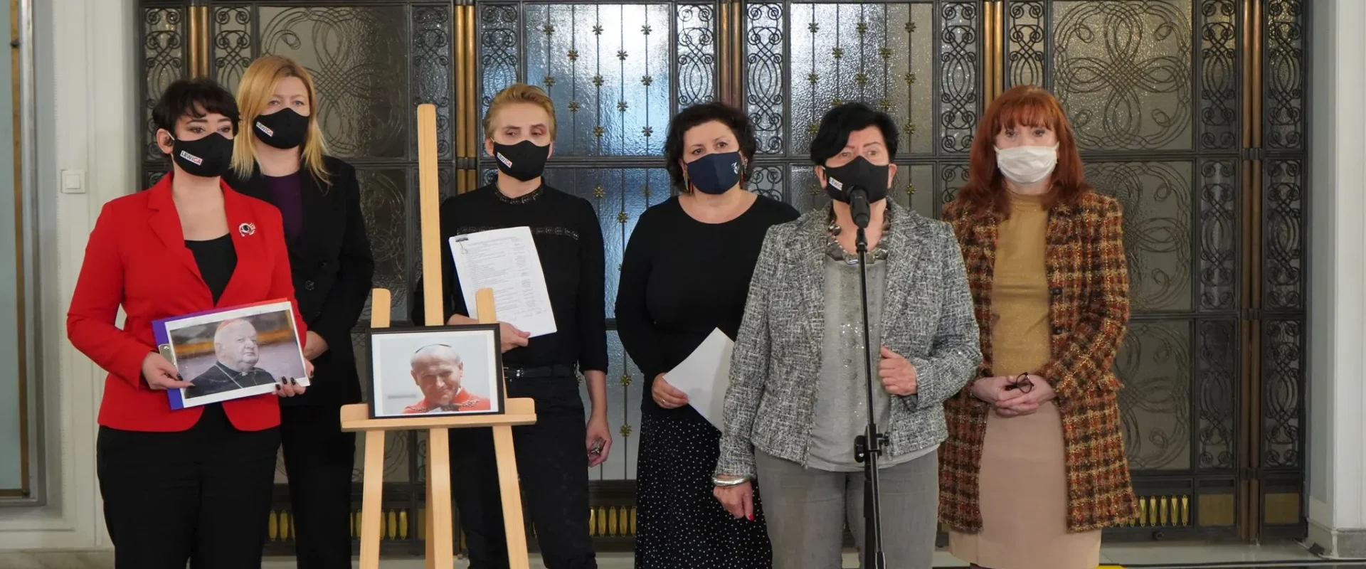Grupa kobiet w maseczkach stoi przy mikrofonie podczas konferencji prasowej, trzymając zdjęcia duchownych na sztaludze, w tle ozdobne drzwi.