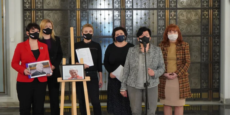 Grupa kobiet w maseczkach stoi przy mikrofonie podczas konferencji prasowej, trzymając zdjęcia duchownych na sztaludze, w tle ozdobne drzwi.