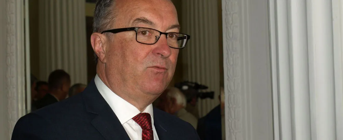 Mężczyzna w granatowym garniturze i czerwonym krawacie, noszący okulary, stoi w wnętrzu z kolumnami.