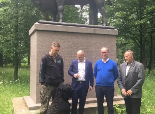 Czterech mężczyzn w różnym wieku stoi przed dużym pomnikiem żubra w leśnym otoczeniu, obok nich siedzi duży czarny pies.