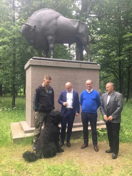 Czterech mężczyzn w różnym wieku stoi przed dużym pomnikiem żubra w leśnym otoczeniu, obok nich siedzi duży czarny pies.