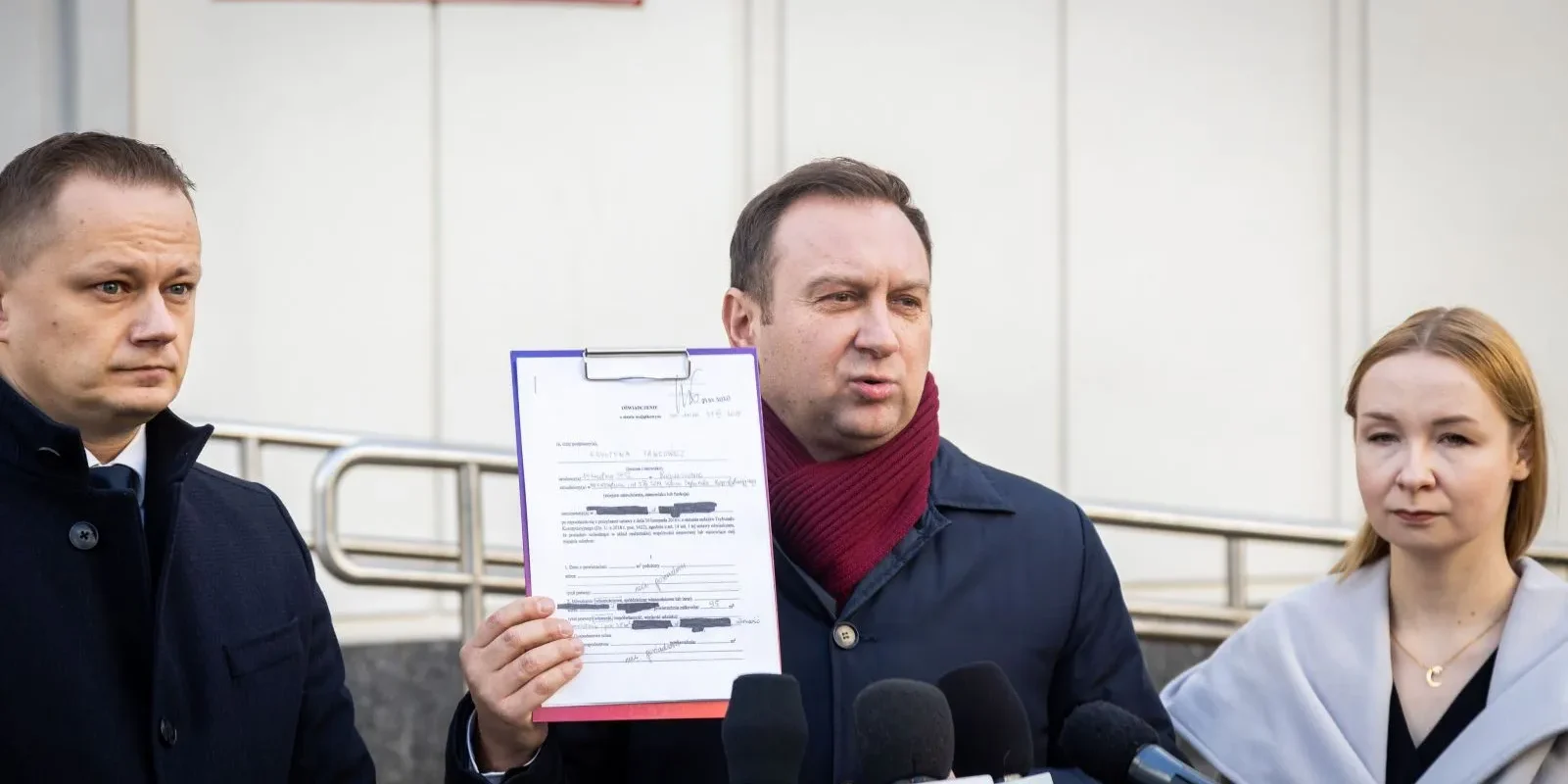 Na konferencji prasowej trzy osoby stoją blisko siebie; mężczyzna w środku prezentuje podpisany dokument, wokół niego ustawione są mikrofony mediów.