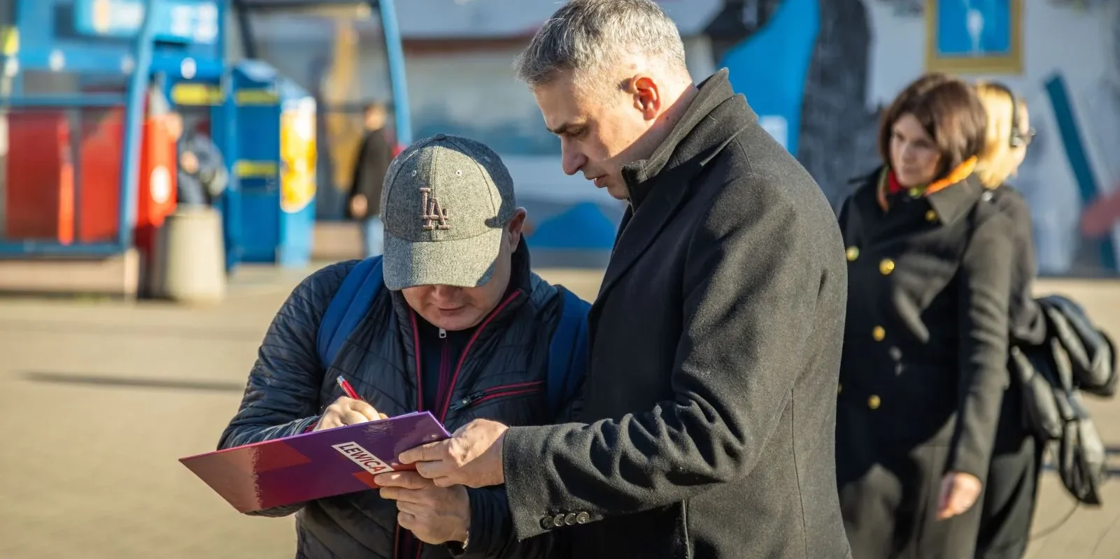Na zdjęciu dwaj mężczyźni rozmawiają i przeglądają dokumenty na tle miejskiego krajobrazu.
