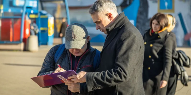 Na zdjęciu dwaj mężczyźni rozmawiają i przeglądają dokumenty na tle miejskiego krajobrazu.