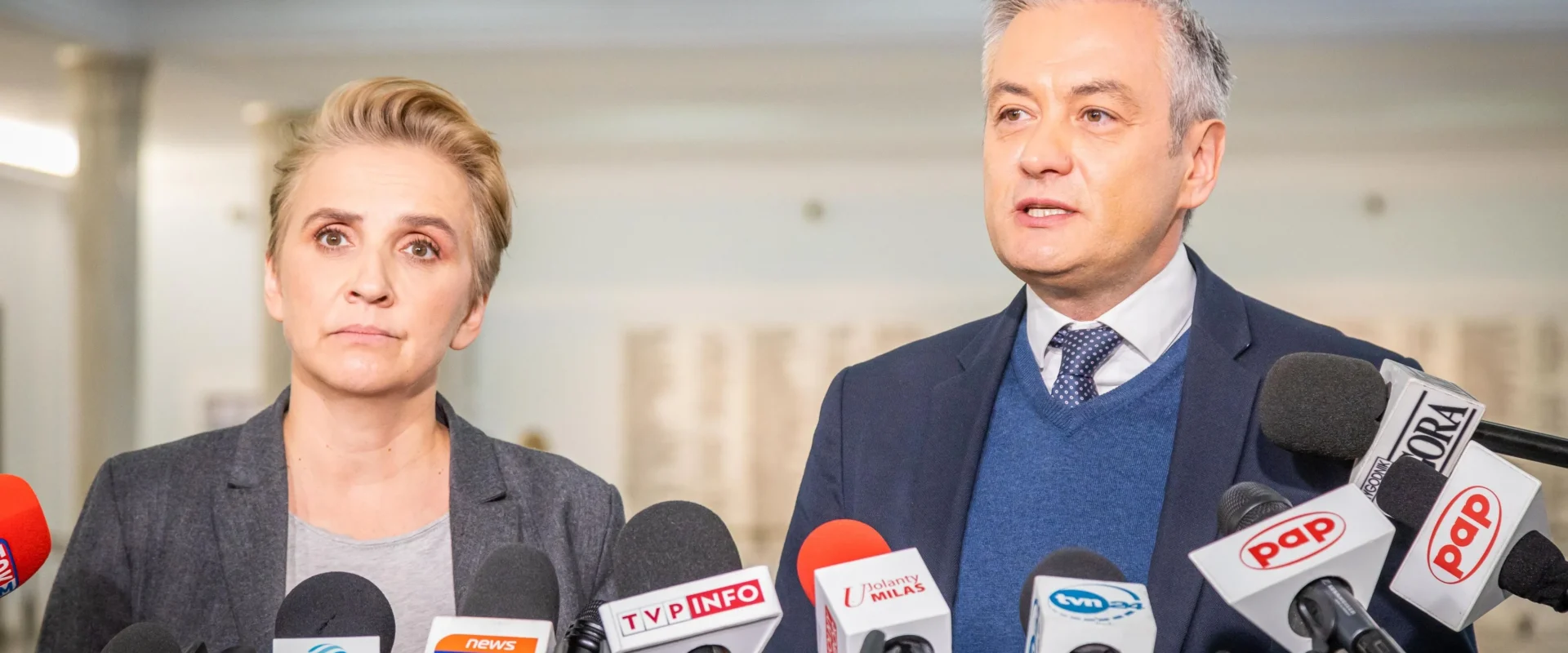 Na zdjęciu dwóch mówców stoi przed mikrofonami podczas konferencji prasowej, prezentując poważne wyrazy twarzy.