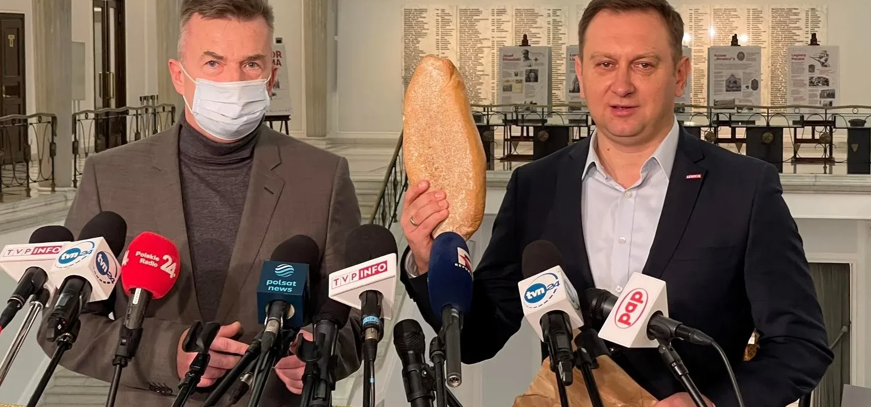 Na zdjęciu dwóch mężczyzn, jeden z nich trzyma bochenek chleba, stoi przed mikrofonami w lokalu publicznym.