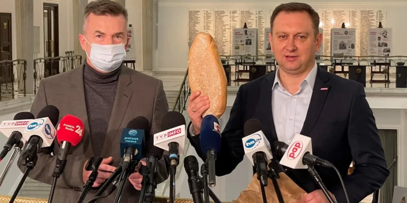 Na zdjęciu dwóch mężczyzn, jeden z nich trzyma bochenek chleba, stoi przed mikrofonami w lokalu publicznym.